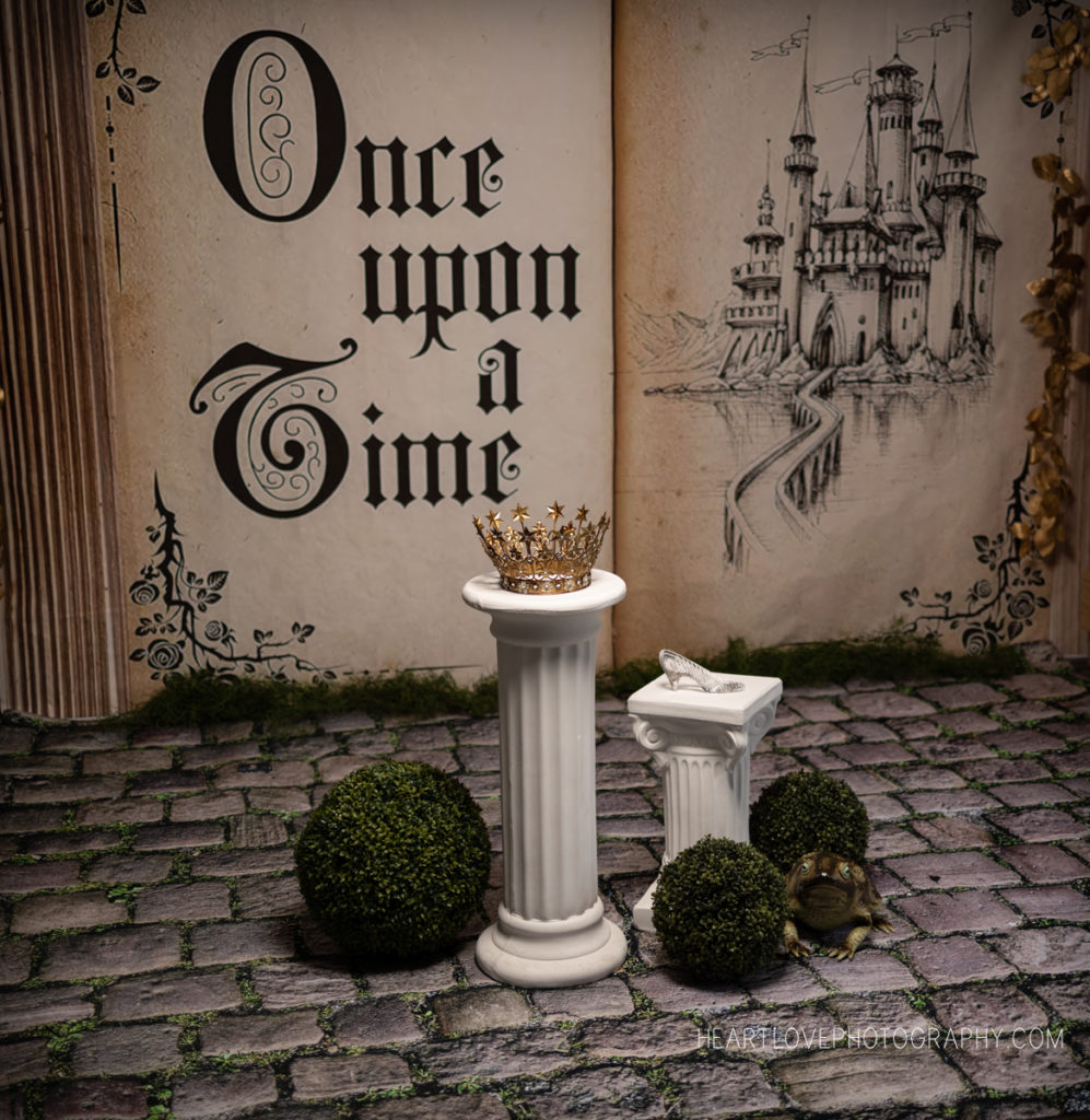 storybook page backdrop reads "once upon a time" with a line work castle, cobblestone floor, white pillars, crown and glass slipper, frog
