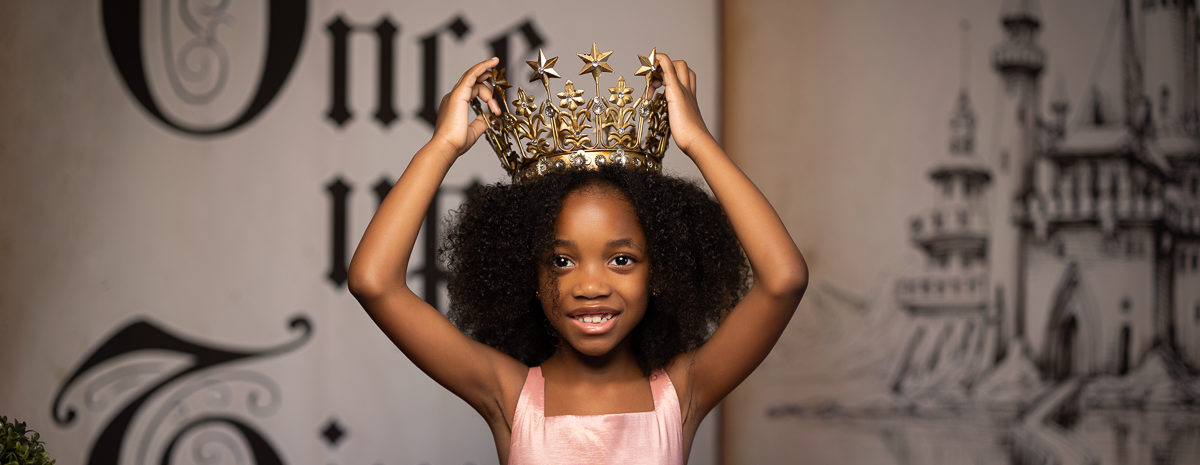Girl with pink dress holding gold crown on a storybook backdrop